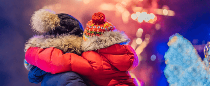 Parent holding child, both dressed in winter clothing, watching fireworks