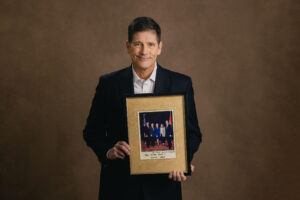 Andy McCain holds a picture frame. The picture in the frame is himself, his father John McCaine, and others.